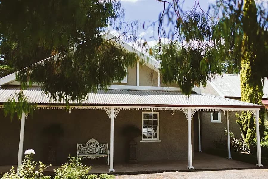 Country house with a shaded veranda beneath leafy trees.