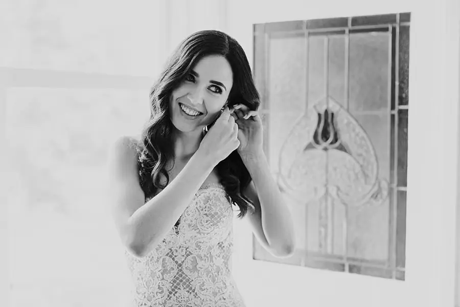 Black-and-white photograph of the smiling bride putting on an earring.