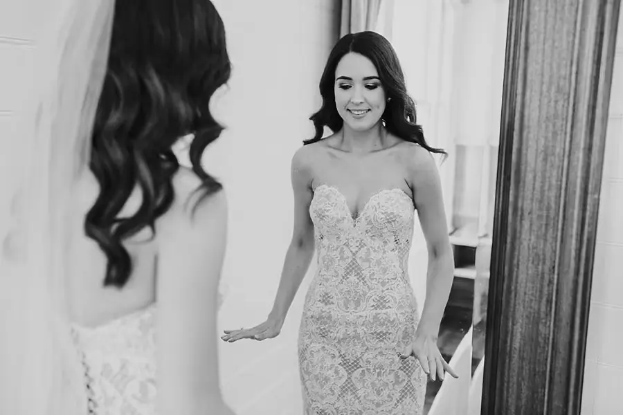 Black-and-white photograph of the bride looking at her lace dress in a mirror.
