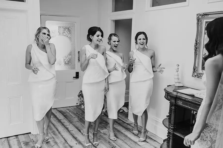 Black-and-white photograph of five women reacting to the bride's wedding dress.