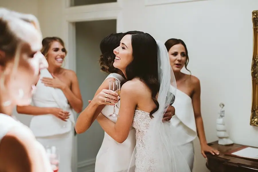 Bride laughing and holding a drinking glass among women in white dresses.