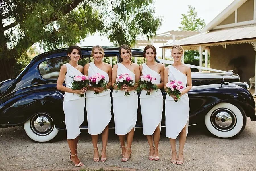Five women in white dresses posing with pink bouquets beside a black vintage car.