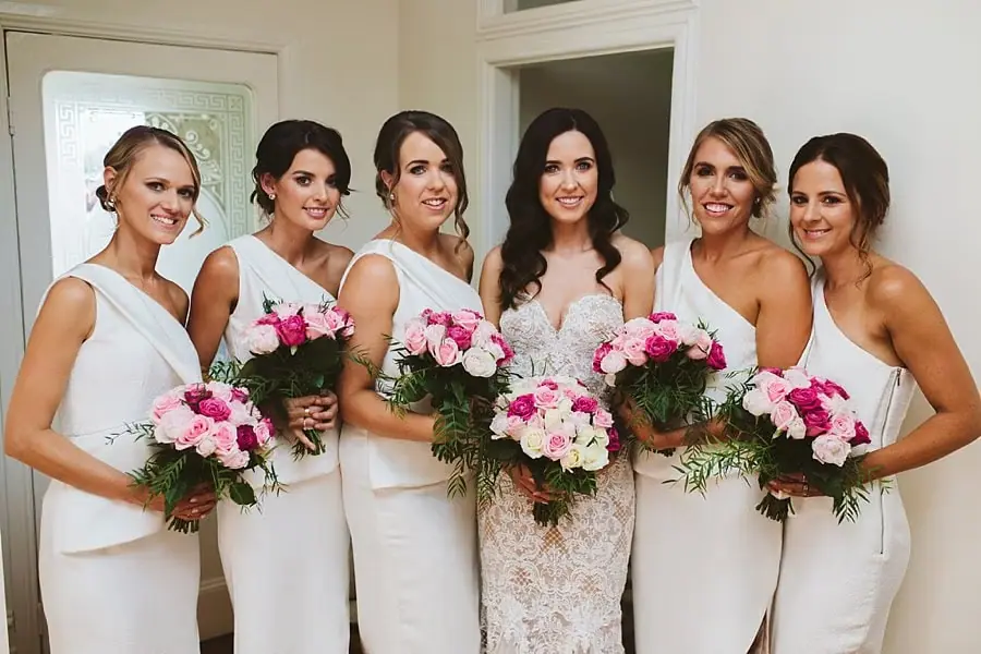 Bride and five women in white dresses smiling together with pink and white bouquets.