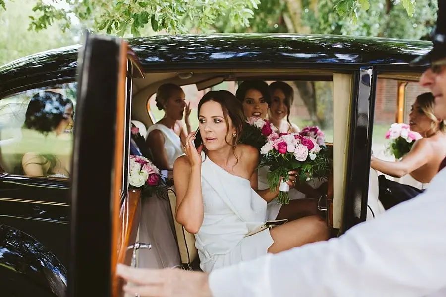 Women in white dresses and pink bouquets sitting inside a black vintage car.