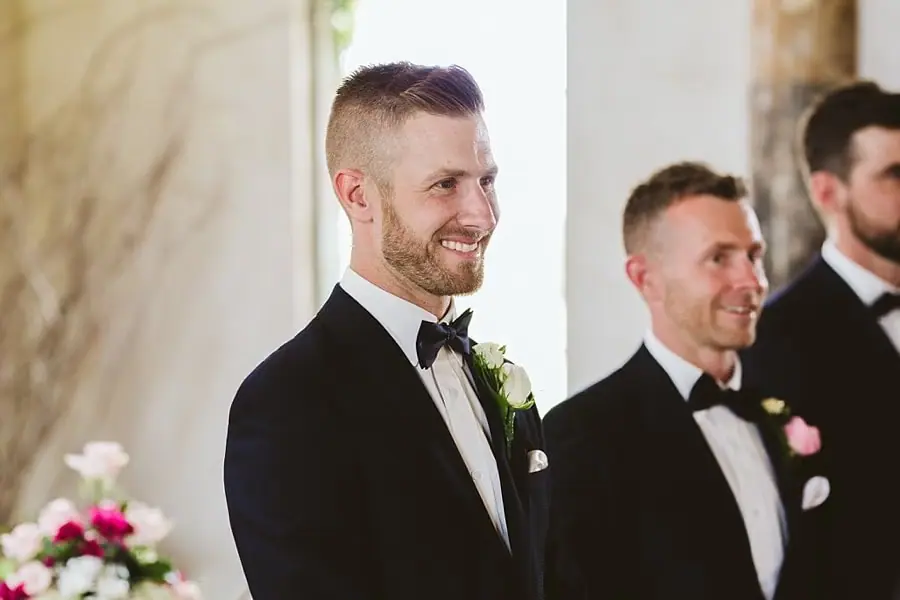 Close portrait of the smiling groom waiting beside his companions in the chapel.