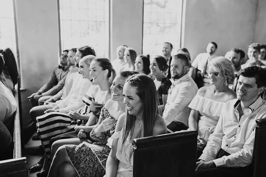 Black-and-white photograph of guests smiling in rows of wooden chapel pews.