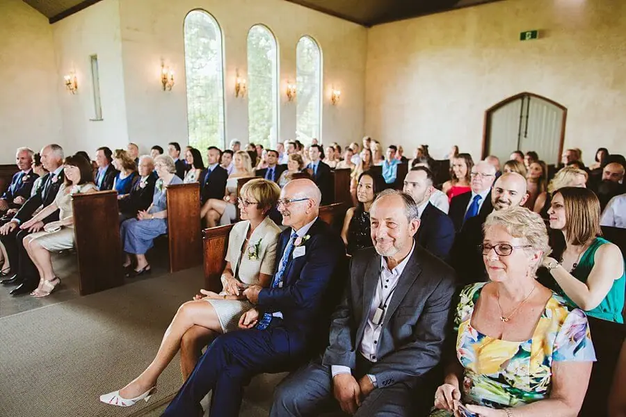 Guests seated in wooden chapel pews beneath tall arched windows.
