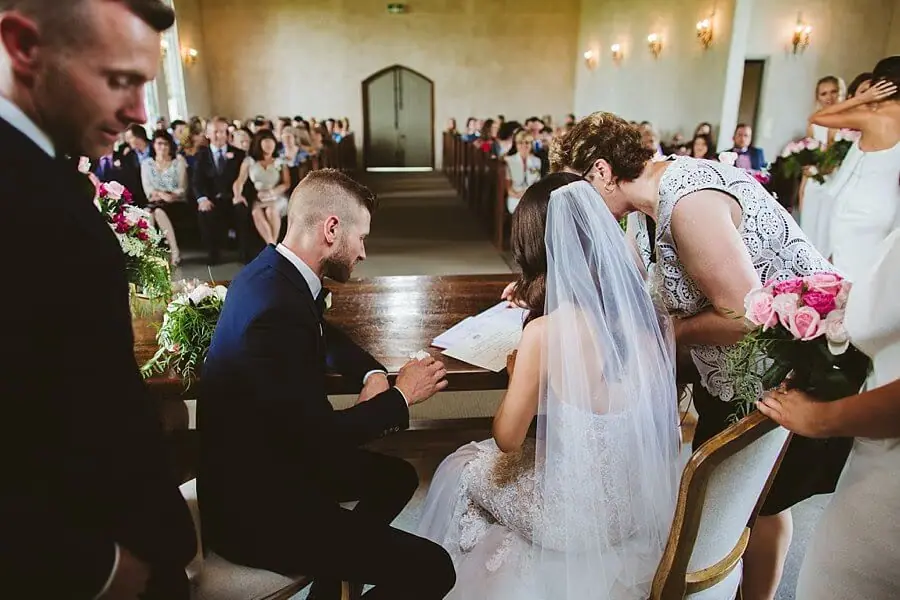 Bride and groom signing paperwork at a table while companions watch.