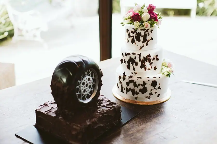 Chocolate tyre-shaped cake beside a three-tier white cake topped with pink flowers.