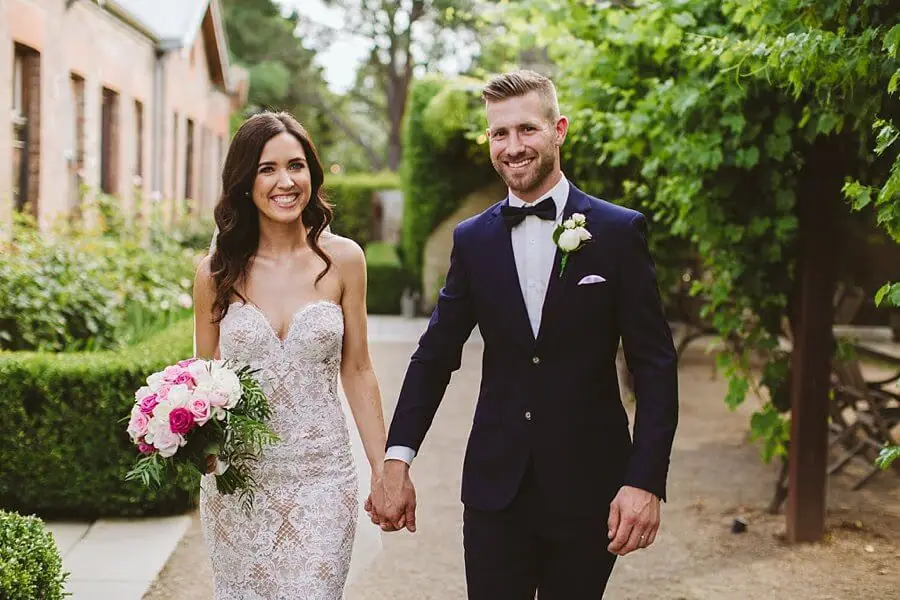 Bride and groom smiling and holding hands on a leafy garden path.