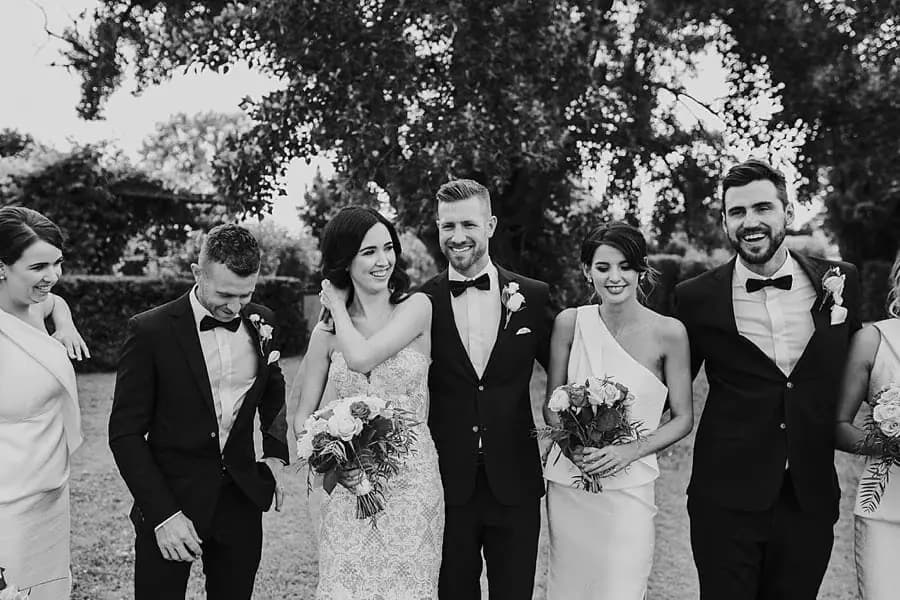 Black-and-white photograph of the couple laughing with their wedding party outdoors.