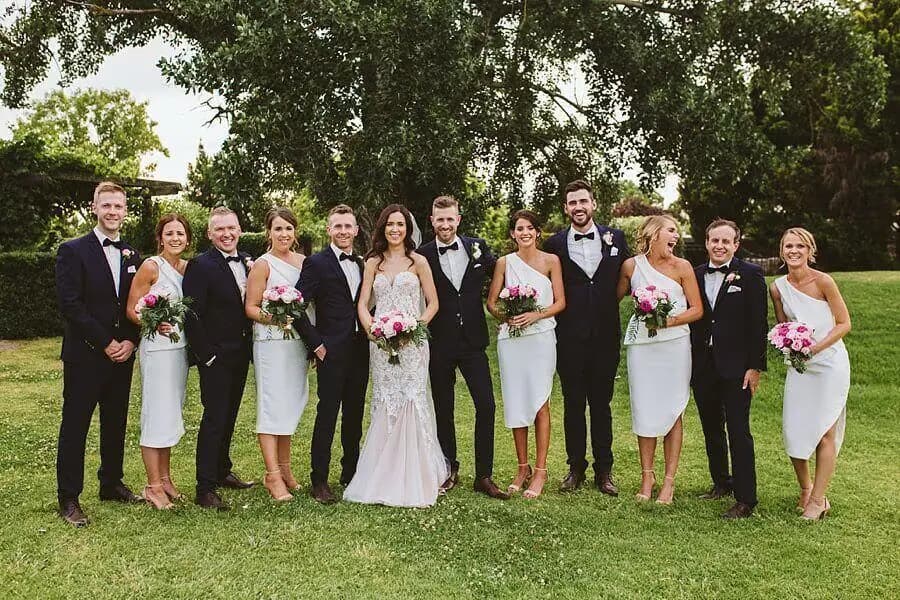 Wedding party lined up on a green lawn beneath large trees.
