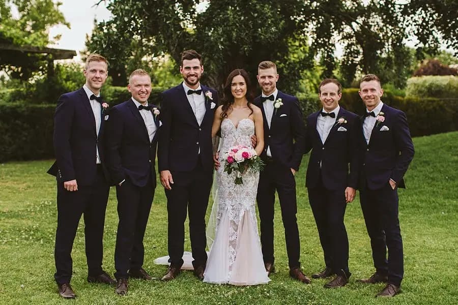 Bride posing with six men in dark suits on a garden lawn.