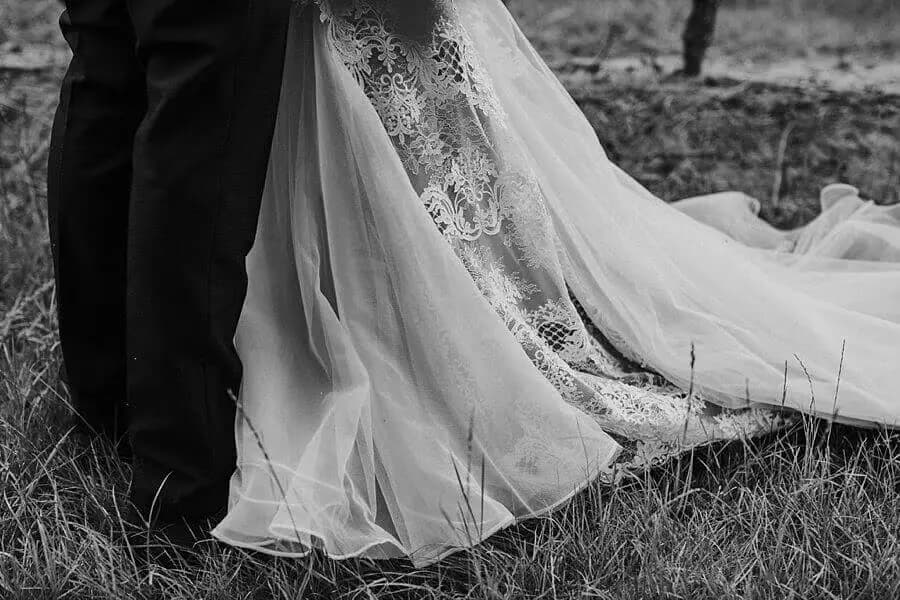 Black-and-white close-up of the bride's lace dress and flowing veil beside the groom's trousers.