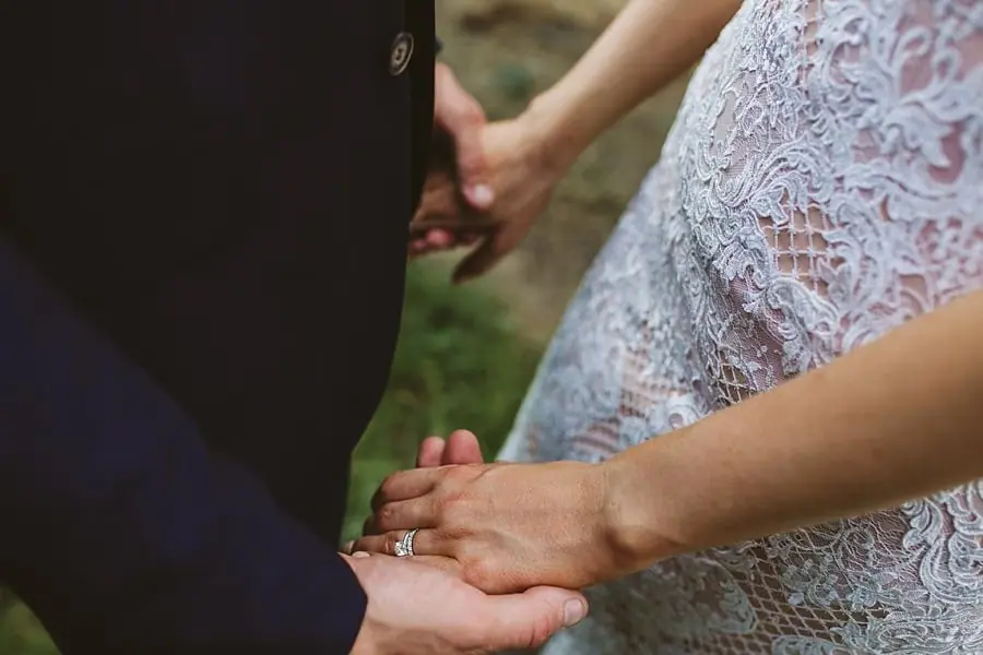 Close-up of the couple holding hands with the bride's ring beside her lace dress.