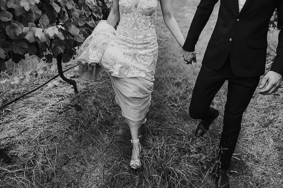 Black-and-white detail of the couple walking hand in hand through a vineyard row.