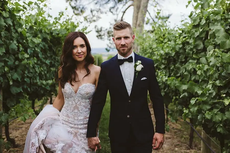 Bride and groom holding hands while walking between leafy vines.
