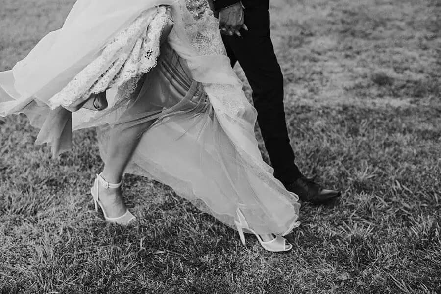 Black-and-white detail of the bride's dress and shoes as the couple walk across grass.