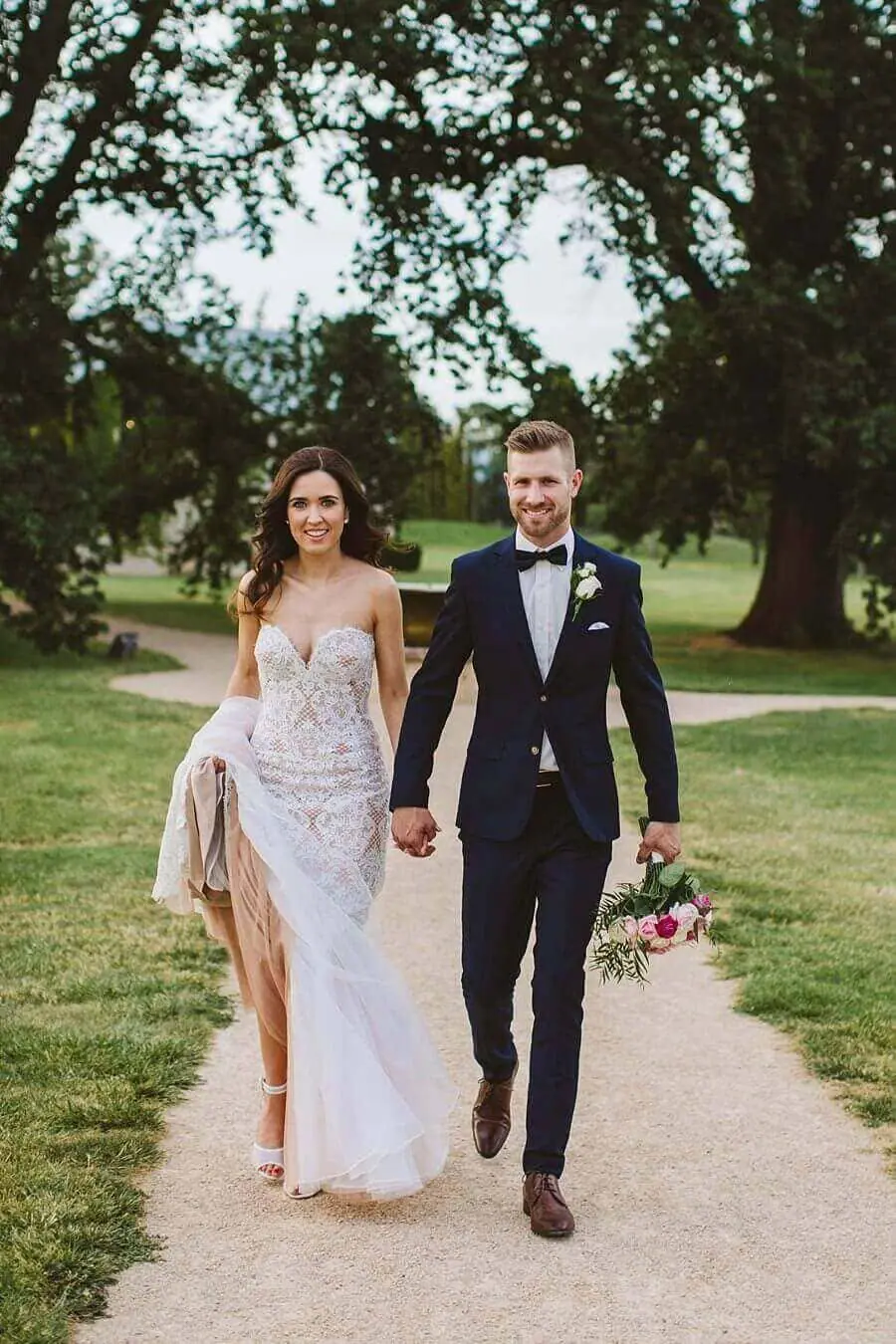 Bride and groom smiling together on a garden path while he carries her bouquet.