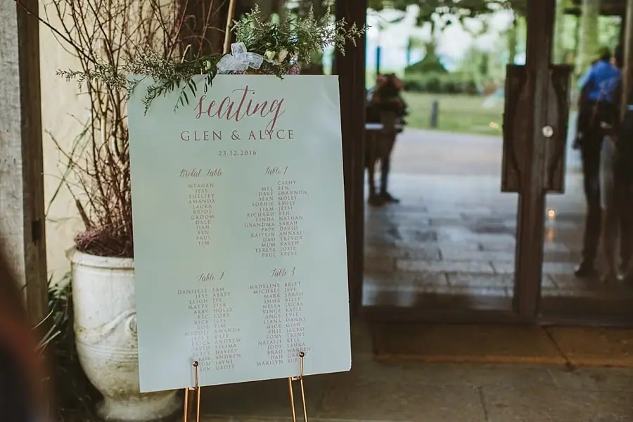 White reception seating chart displayed on an easel beside glass doors.