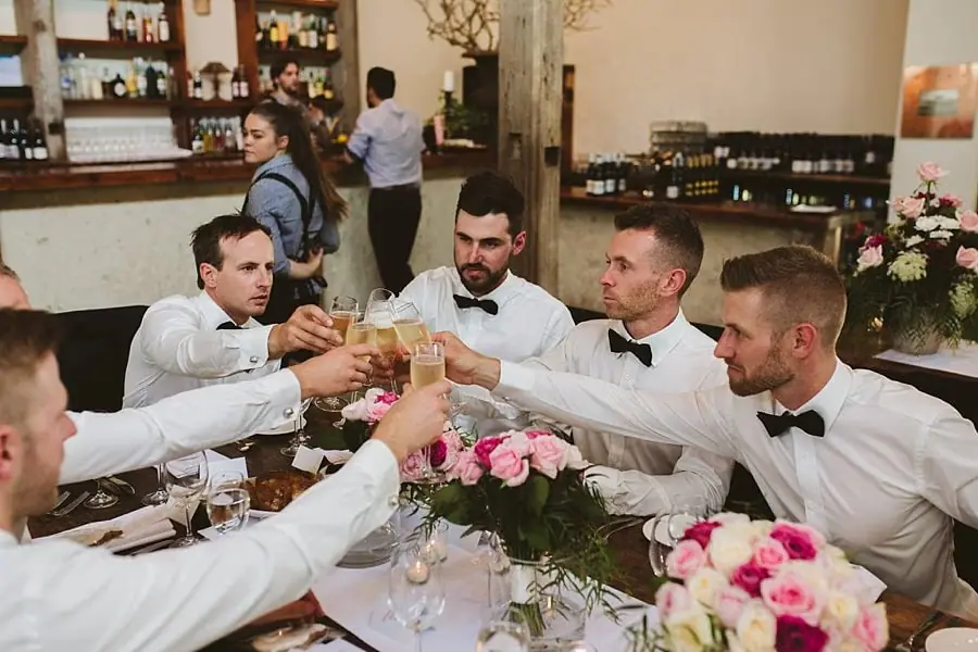 Men clinking drinking glasses across a flower-decorated reception table.