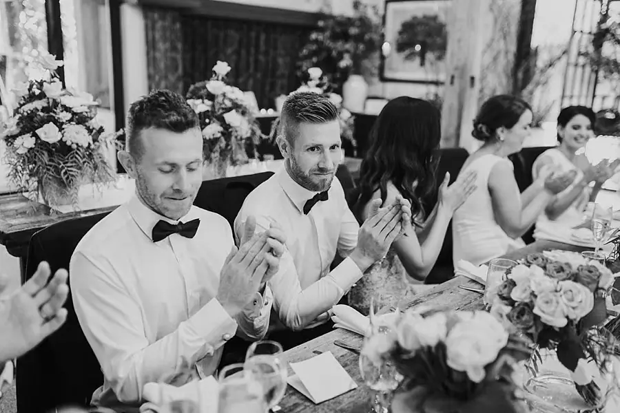 Black-and-white photograph of wedding party members applauding at a reception table.