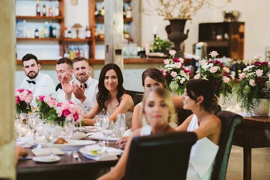 Bride and groom listening alongside their wedding party at a flower-decorated table.