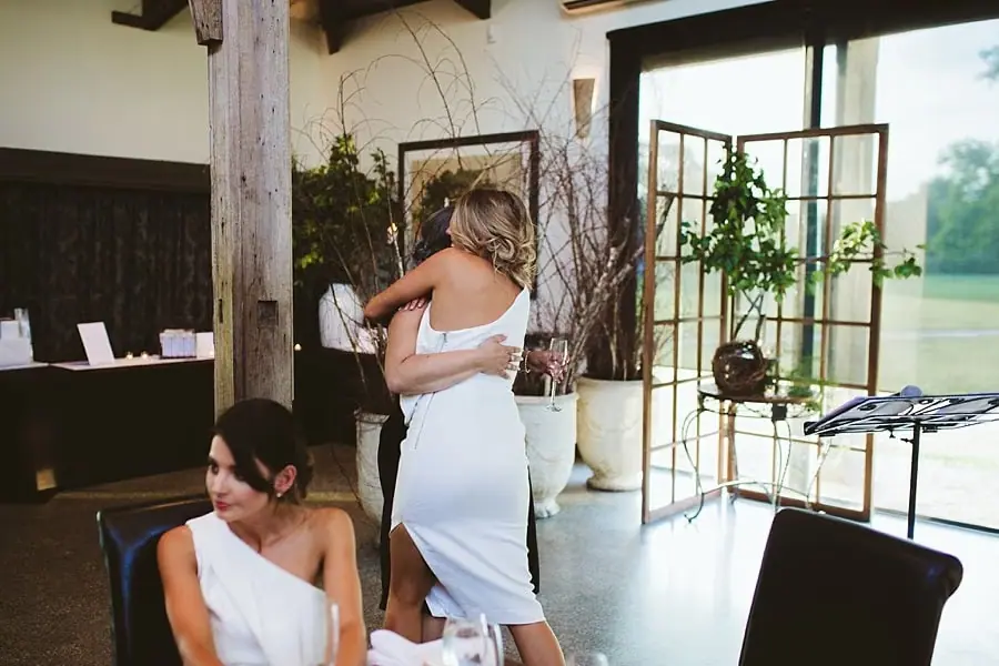 Woman in a white dress embracing a companion beside a timber pillar.