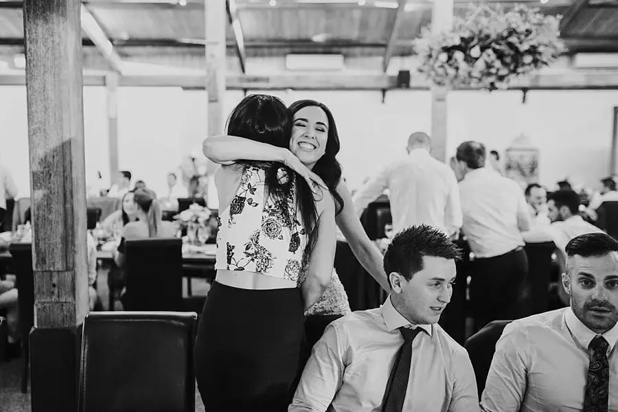 Black-and-white photograph of the bride hugging a guest beside a reception table.