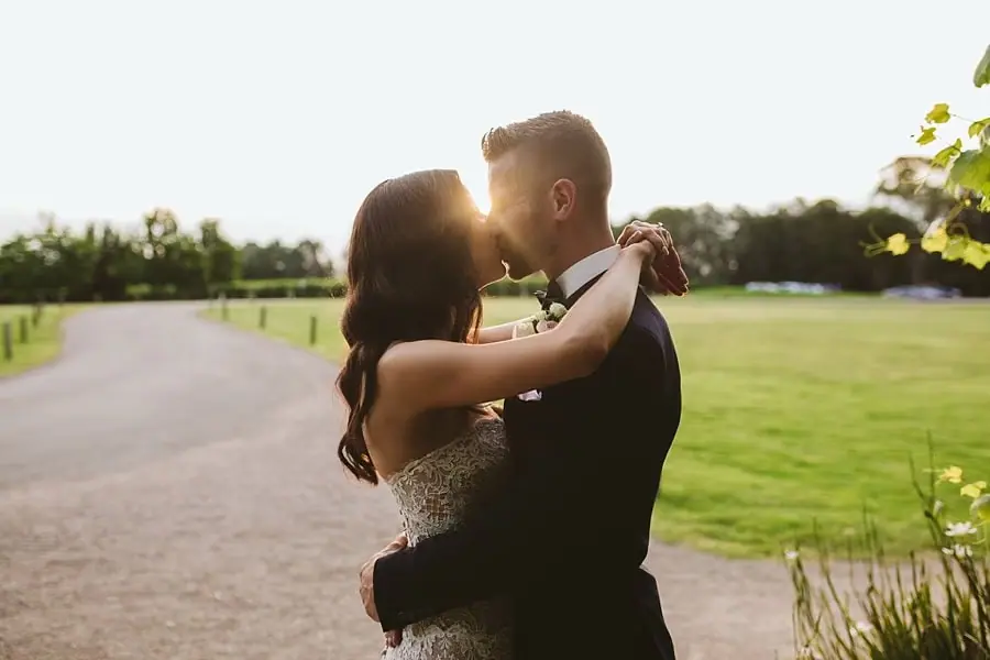 Couple embracing beside a garden driveway in warm late-afternoon light.
