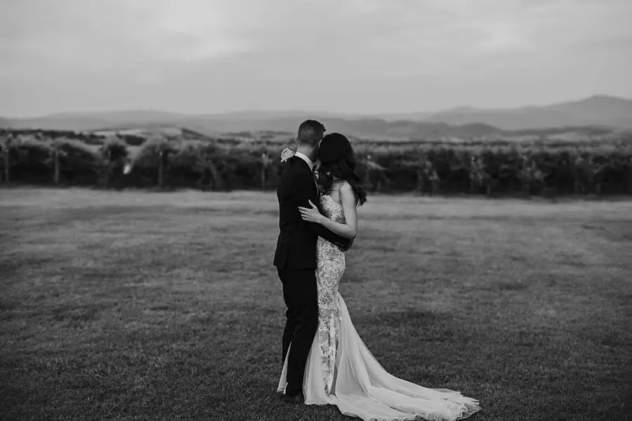 Black-and-white portrait of the couple embracing beside vineyard rows.