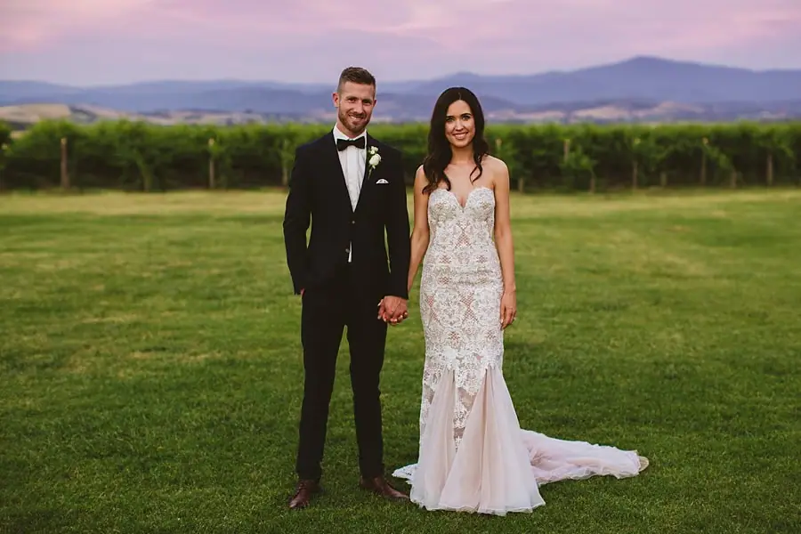 Bride and groom holding hands on a lawn with mountains beneath a pink sky.