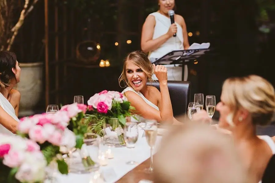 Woman laughing at the reception table while another woman gives a microphone speech.