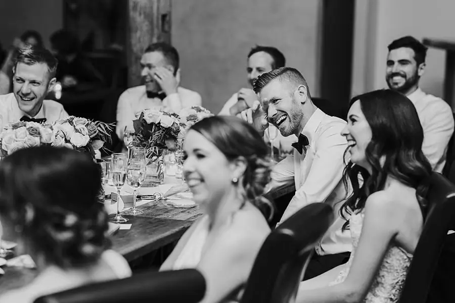 Black-and-white photograph of the couple and their wedding party laughing at a reception table.