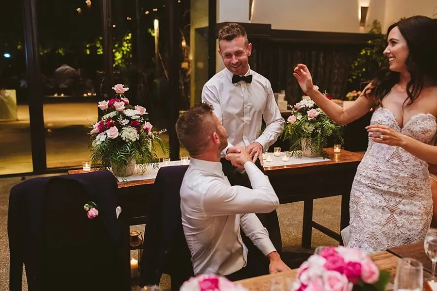 Bride and two men sharing a playful moment beside the reception table.