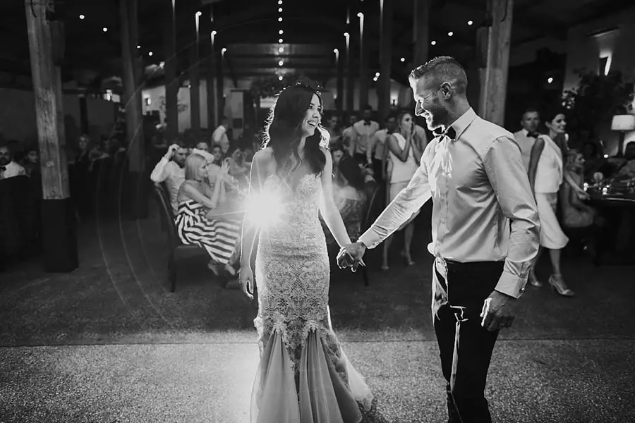 Black-and-white photograph of the couple holding hands on the dance floor with bright backlighting.