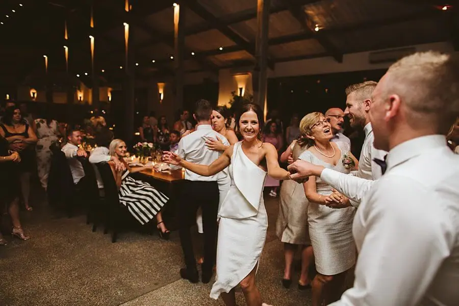 Woman in a white dress raising her arms as guests dance around her.