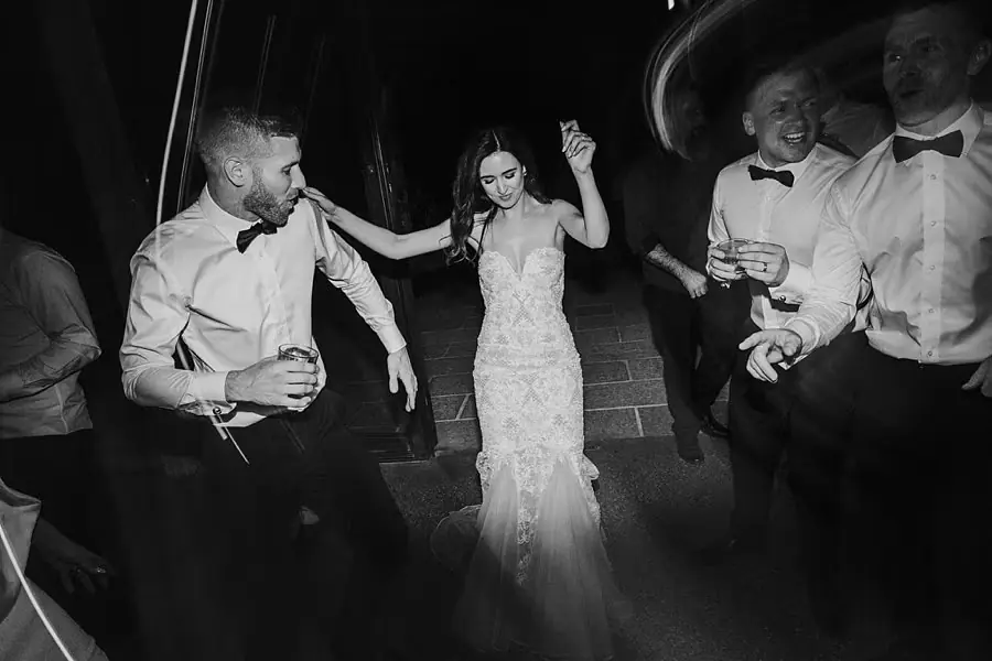 Black-and-white photograph of the bride dancing among men holding drinks.
