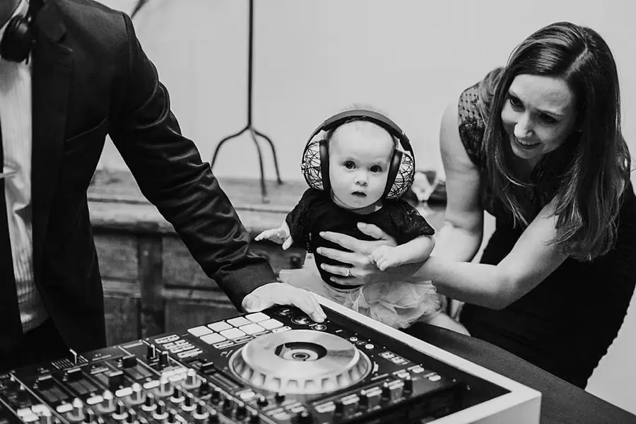 Black-and-white photograph of a woman holding a baby wearing ear defenders beside DJ equipment.