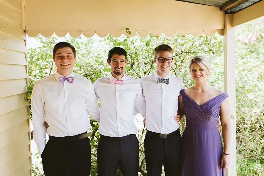 Three men in white shirts and bow ties posing with a woman on a verandah