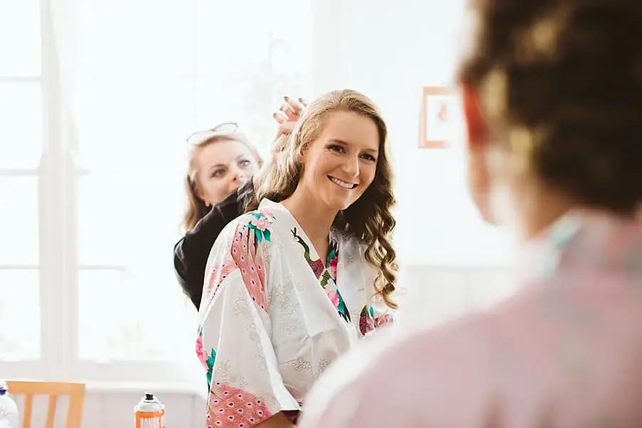 Bride smiling as a stylist arranges her hair beside a mirror