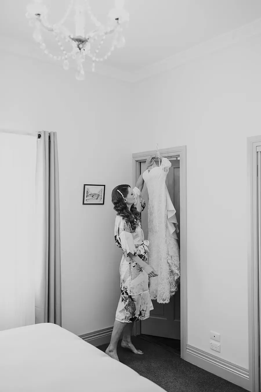 Black-and-white photograph of the bride reaching toward her hanging wedding dress