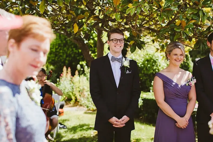 Groom smiling beside a woman in a purple dress beneath green foliage