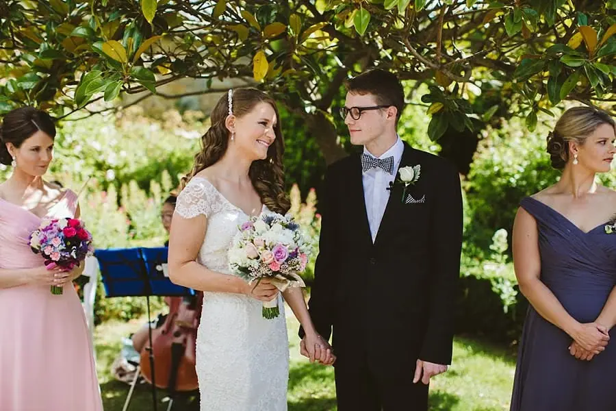 Bride and groom holding hands beneath a leafy tree with their wedding party