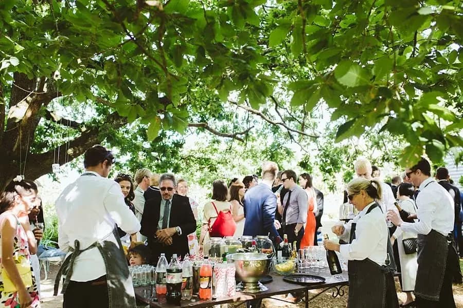 Guests gathering around outdoor drinks tables beneath a leafy tree