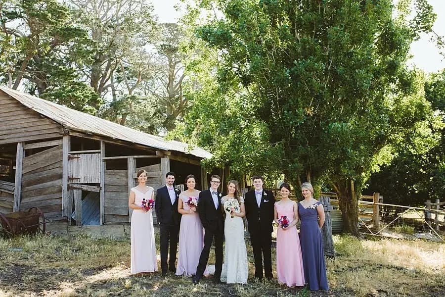 Wedding party standing beside a weathered wooden shed in the shade of trees