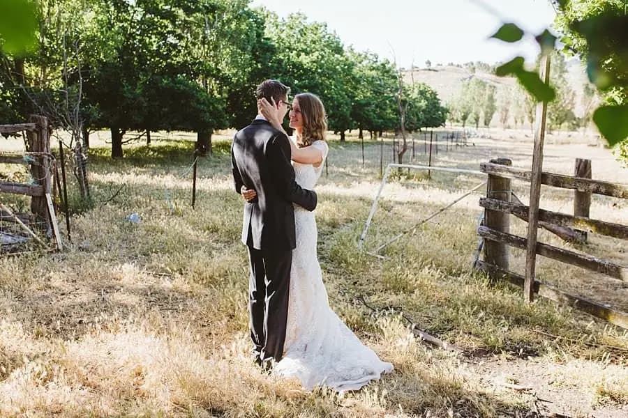 Bride and groom embracing beside a timber fence and orchard trees
