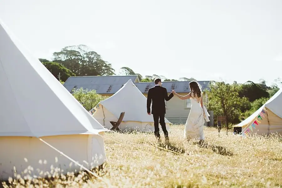 Bride and groom walking away together between bell tents in a grassy field