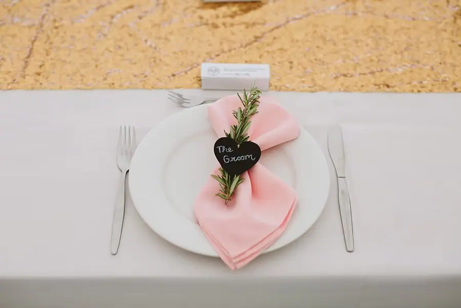Place setting with a pink napkin, rosemary sprig and heart-shaped groom label