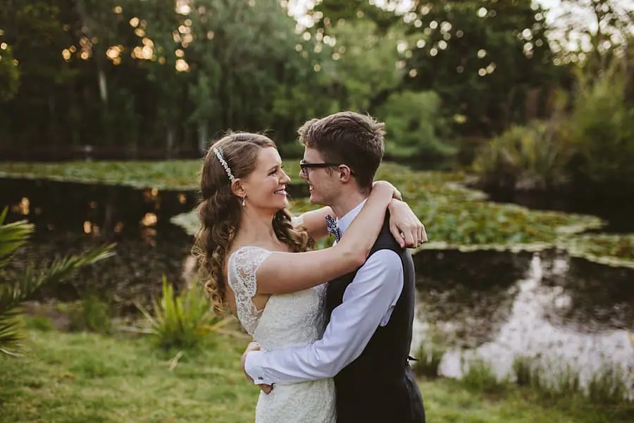 Bride and groom embracing beside a pond in warm evening light